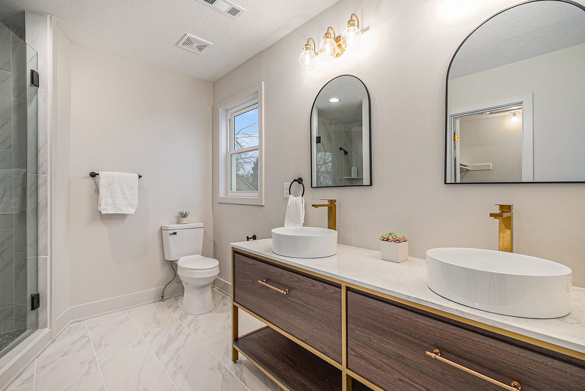 After: primary bath with a wood-and-gold double vanity, vessel sinks, arched mirrors, and a marble shower
