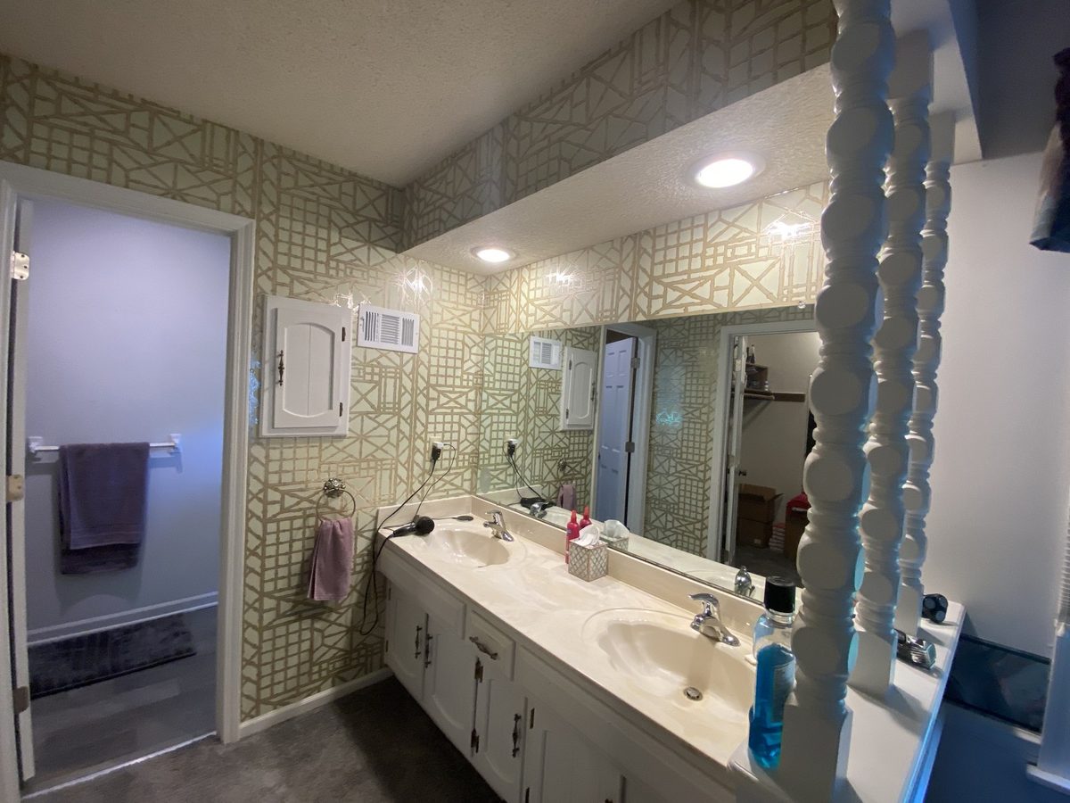Before: primary bath with a double vanity, trellis wallpaper, and a cultured-marble countertop