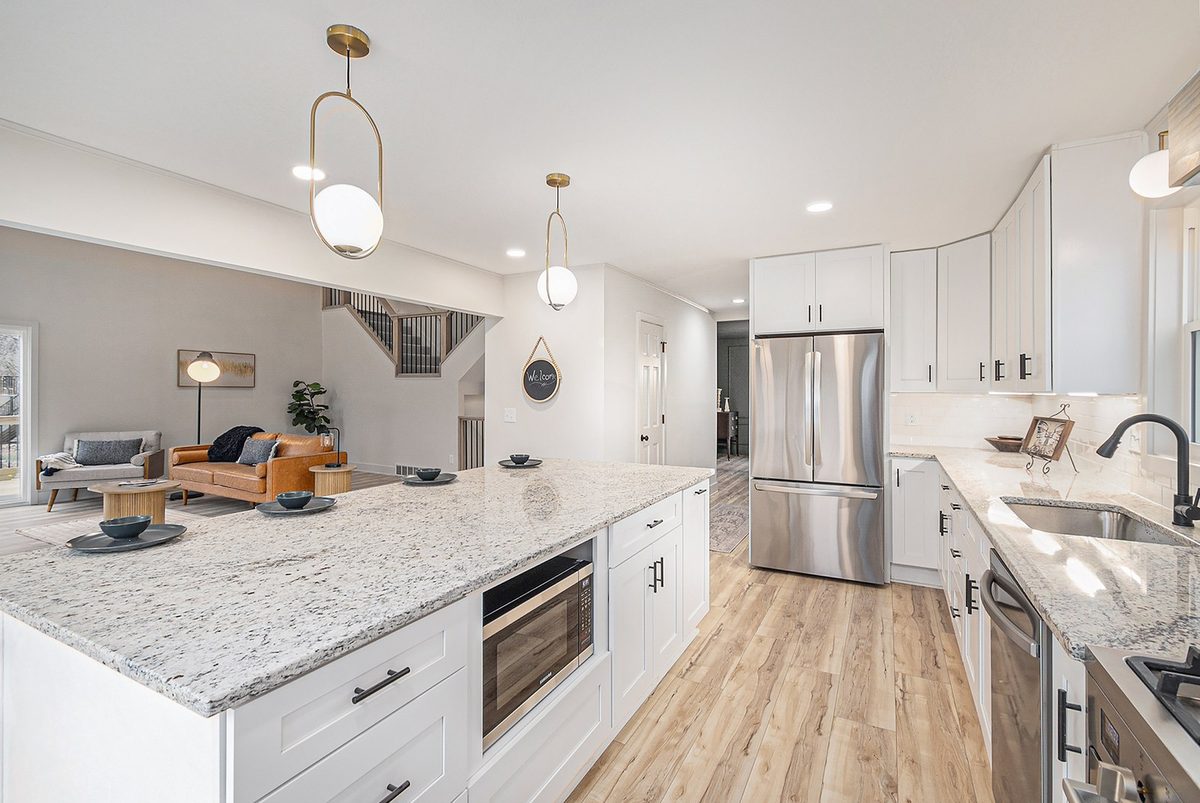 After: bright white kitchen with a granite island, stainless appliances, and wood-look floors open to the living room